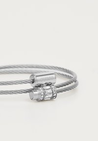 Bracelet en argent avec un design en fil torsadé, fermoir métallique gravé "GUESS" et une bande décorative ornée de strass transparents.