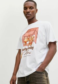 Tânăr îmbrăcat cu un tricou alb pe care se află o grafică cu patru figuri de turta dulce stând sub un acoperiș de casă și inscripția "Biscuiți Veseli".