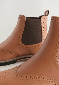 Bruine leren Chelsea-laarzen met brogue-perforaties en zwarte elastische zijpanelen, close-up weergegeven tegen een lichte achtergrond.