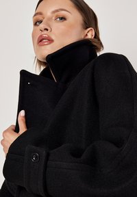 Cappotto in misto lana nero con colletto alto, polsini abbottonati e silhouette strutturata. Caratterizzato da una finitura del tessuto liscia e texturizzata.