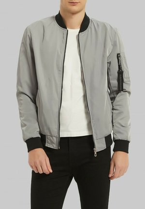 Veste bomber grise avec fermeture éclair à l'avant, col et poignets côtelés noirs, poches latérales et poche zippée sur la manche gauche.