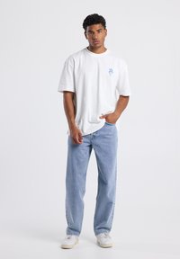 White cotton t-shirt, relaxed fit with short sleeves, featuring blue graphic detail on the left chest. Light blue denim jeans, straight leg.