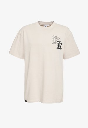 Beżowy bawełniany T-shirt z krótkim rękawem, z czarnym stylizowanym nadrukiem litery "K" na lewej piersi oraz małą metką marki przy dolnym szwie.