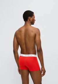 Κόκκινα ανδρικά μποξεράκια με λευκή ελαστική ζώνη μέσης που φέρει το λογότυπο "TOMMY HILFIGER". Λείο υλικό, εφαρμοστή σχεδίαση, κομμένα λίγο πάνω από τη μέση του μηρού.