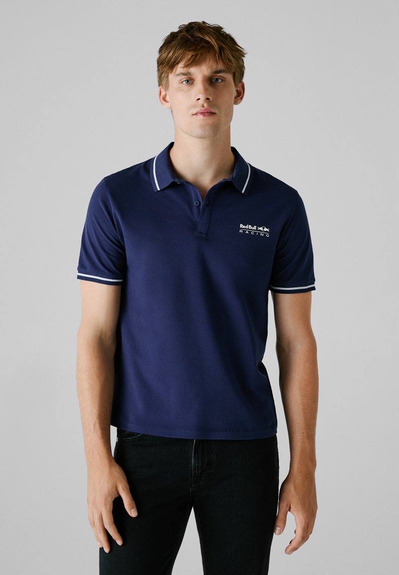 Marineblauwe polo met korte mouwen, witte accenten op de kraag en mouwen, voorzien van een logo op de linkerborst, gemaakt van een gladde stof.