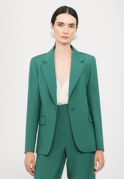 Teal blazer med snipper og enkelt knappelukking, med en brystlomme og to sidelommer. Bært over en lys beige topp.