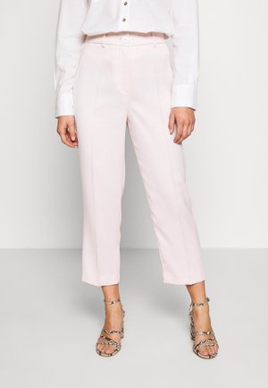 Femme portant un pantalon court rose clair, une blouse blanche boutonnée et des sandales à talons imprimées serpent, debout devant un fond uni.