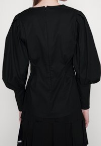 Czarna bluzka z bufkami na długich rękawach, okrągłym dekoltem i zapięciem na zamek z tyłu. Gładka tekstura z wyraźnymi szwami.