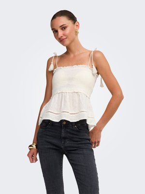 Jeune femme portant un haut blanc sans manches à volants avec des bretelles à nouer et un jean noir, posant avec une main sur la hanche sur un fond uni.