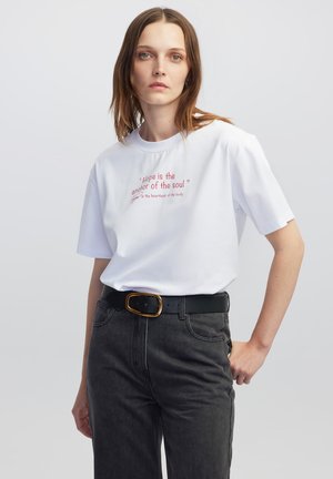Witte katoenen t-shirt met korte mouwen met rode tekst. Gecombineerd met donkergrijze jeans en een zwarte riem met een gouden gesp.