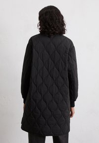 Veste noire matelassée avec un motif texturé de courbes superposées, manches longues et poignets côtelés, présentant un design mi-long.