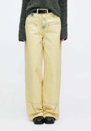 Vide ben, gule dongeribukser med synlige sømmer, falmede detaljer og røde metallknapper. Brukt med en grå teksturert genser og svart belte.