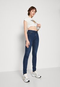 Jeans skinny azul escuro com uma cintura alta, combinados com uma t-shirt cropped branca com um design gráfico. O modelo usa tênis brancos e amarelos.