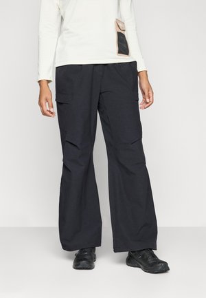Schwarze Cargo-Hosen mit weiter Beinform, strukturiertem Stoff, mehreren Seitentaschen und lockerer Passform. Getragen mit schwarzem Schuhwerk.