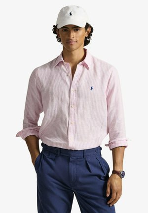 Jeune homme aux cheveux bouclés portant une casquette blanche, une chemise rose clair à rayures et boutons, un pantalon bleu marine et une montre-bracelet.