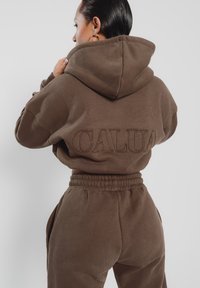 Brauner Hoodie mit "CALUA" auf dem Rücken gestickt, ausgestattet mit einer Kapuze mit Kordelzug und lockerer Passform. Kombiniert mit passenden Hosen mit elastischem Bund.