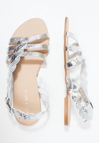 Litte sandaalit, joissa on metallinhohtoiset hopeiset keinonahkaremmit, kermaisen värinen pohja ja puoliksi punottu yksityiskohta. Varustettu säädettävällä nilkkaremmillä ja sileällä pohjalla.