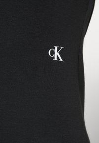 Haut en coton noir présentant un gros plan de la texture du tissu avec un petit logo "cK" blanc sur le côté gauche. Design simple, sans embellissements.