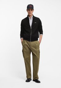 Άνδρας που φοράει μαύρο πουλόβερ με φερμουάρ, ριγέ πουκάμισο, χακί παντελόνι cargo, μαύρα μποτάκια και μαύρο καπέλο με κόκκινο λογότυπο, στέκεται με τα χέρια στις τσέπες.