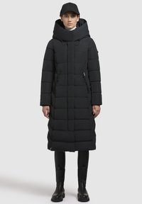 Cappotto piumino nero con colletto alto e cappuccio, caratterizzato da pannelli trapuntati, tasche con zip frontali e silhouette dritta. Abbinato a stivali neri.
