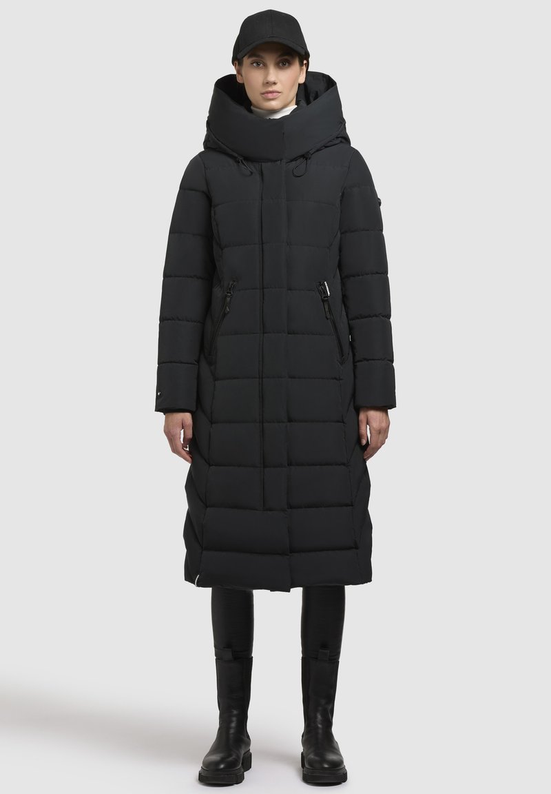 Cappotto piumino nero con colletto alto e cappuccio, caratterizzato da pannelli trapuntati, tasche con zip frontali e silhouette dritta. Abbinato a stivali neri.