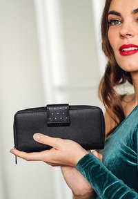 Černá zipová peněženka vyrobená z hladké kůže, s texturovaným černým zámkem a malými nýty. Držena v ruce na neutrálním pozadí.