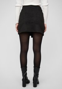 Zwarte suède minirok met een gebogen zoom, gecombineerd met doorschijnende zwarte panty's en knielange zwarte laarzen met hak. Achteraanzicht weergegeven.
