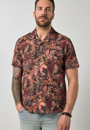 Homme avec barbe et tatouage portant une chemise à manches courtes avec un motif de feuilles tropicales rouges et brunes et un jean bleu clair.
