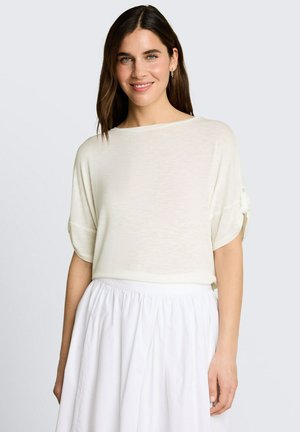 Glunderende vrouw met stijlbruin haar, die een los wit shirt met korte mouwen en een witte geplooide rok draagt tegen een lichte achtergrond.