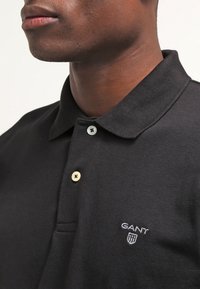 Musta polo-paita, joka on valmistettu kim 활cältä kankaasta, jossa on klassinen kaulus, kahden napin napitus kultaisilla ja mustilla napeilla sekä brodeerattu logo.