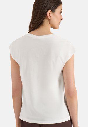 Femme aux longs cheveux bruns portant un haut blanc uni à manches courtes, vue de dos sur un fond blanc.