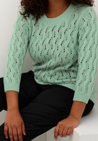 Pull tricoté vert menthe à coupe ample, avec un motif ajouré et des manches trois-quarts. Associé à un pantalon noir.