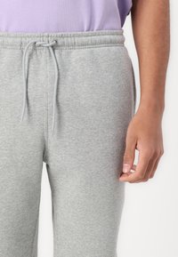 Γκρι παντελόνι φούτερ με ελαστική μέση και κορδόνι, με απαλή υφή και εφαρμοστά πόδια. Χέρι στο κάδρο, φοράει ανοιχτόμωβ πουκάμισο.