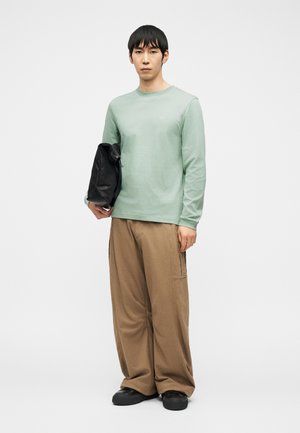 Homme portant une chemise à manches longues vert clair, un pantalon large marron, des chaussures noires, tenant une veste noire pliée sur fond blanc.