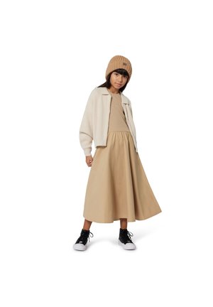 Bonnet beige en tricot, veste courte blanche et duveteuse, haut côtelé beige et longue jupe beige. Des baskets noires à semelles blanches complètent la tenue.
