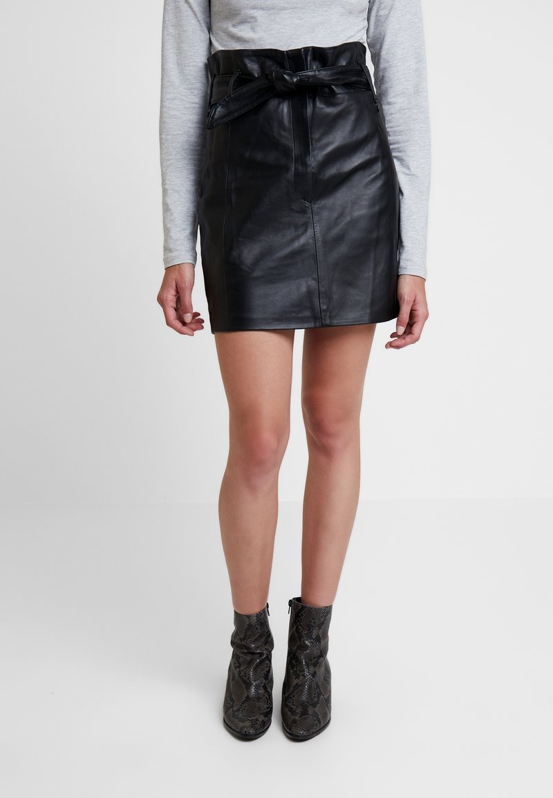 Femme portant une jupe en cuir noire taille haute avec ceinture nouée, un haut gris à manches longues et des bottines grises à motifs, debout sur un sol blanc.
