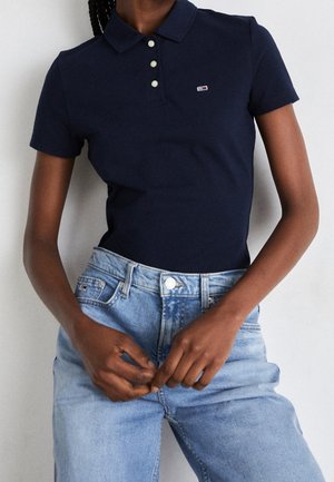 Osoba ubrana w granatową krótką koszulkę polo z małym logo i jasnoniebieskie dżinsy z wysokim stanem, poprawiająca pasek w spodniach.