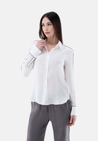 White long-sleeve shirt in smooth fabric with black piping along the seams and buttons. Slightly curved hem, paired with gray trousers.