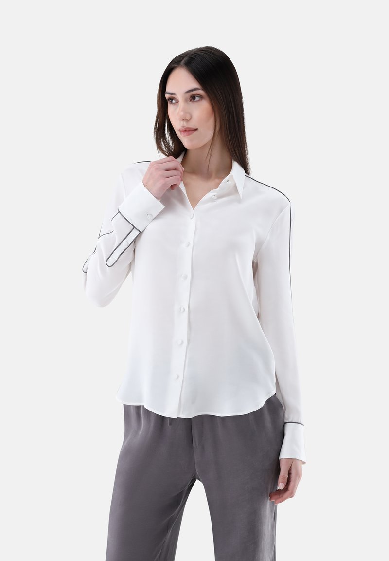 White long-sleeve shirt in smooth fabric with black piping along the seams and buttons. Slightly curved hem, paired with gray trousers.