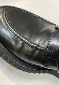 Gros plan d'une chaussure en cuir noire avec des coutures détaillées et une semelle en caoutchouc texturée sur un fond clair.
