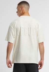 Kremkrāsas īsām piedurknēm krekls ar teksturētu "PEQUS" logo aizmugurē. Audums izskatās mīksts, ar brīvu piegriezumu un sānu šķelumiem.