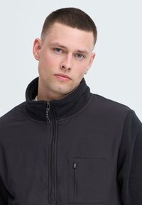 Schwarze Zip-Jacke mit hohem Kragen, aus strukturiertem Stoff, mit seitlicher Tasche mit Reißverschluss und schlichtem Design. Glatter und warmer Stoff.