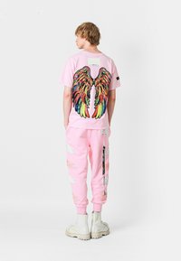 Sudadera rosa con un gráfico colorido de alas en la parte posterior; pantalones de chándal rosa a juego con patrones de plumas blancas, combinados con botas blancas.