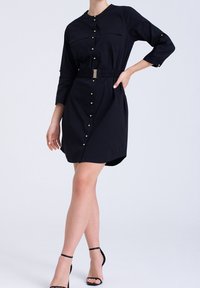 Svart skjortklänning med långa ärmar, knappfront, två bröstfickor, elastisk midja med guldaccent och en böjd fåll, parat med svarta sandaler.