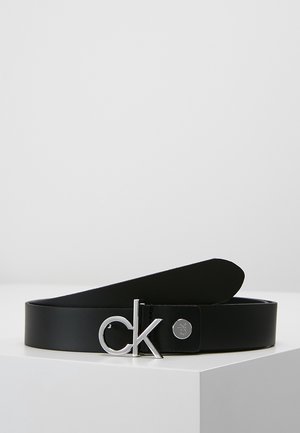 Černý kožený pásek se sponou ve tvaru stříbrného loga "ck", stočený na bílém povrchu na jednoduchém světle šedém pozadí.