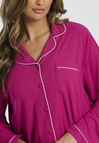 Top de pijama fucsia con textura acanalada, ribete blanco y un bolsillo en el pecho. Presenta un cierre frontal con botones y mangas largas.