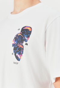 Balta T-krekls ar trim saliktiem neona krāsas skeletiem un gotiskajiem burtiem "O," "R," un "G" ar tekstu "Worldwide Originals" zem grafikas.