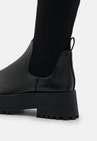 Svart läder ankelboots med en texturerad ribbad sockdel, accentuerad av en dragflik och en tjock, robust sula för ökat grepp.