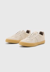Beige Wildledersneaker mit strukturiertem Obermaterial, Schnürverschluss und einer massiven Gummisohle. Verfügt über subtile geometrische Muster an den Seiten.