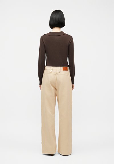 Γυναίκα με κοντά μαύρα μαλλιά φοράει ένα μακρυμάνικο τοπ σκούρου καφέ χρώματος και μπεζ παντελόνι με φαρδιά μπατζάκια, κοιτάζοντας προς τα πίσω σε απλό λευκό φόντο.